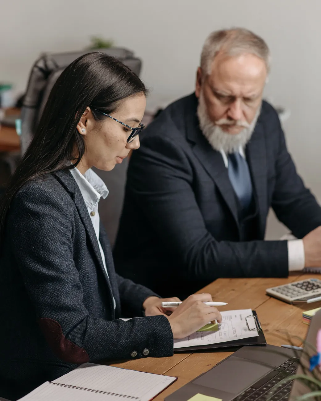 A senior chartered accountant and a colleague reviewing a client's figures on a clipboard, with a calculator and laptop on the desk